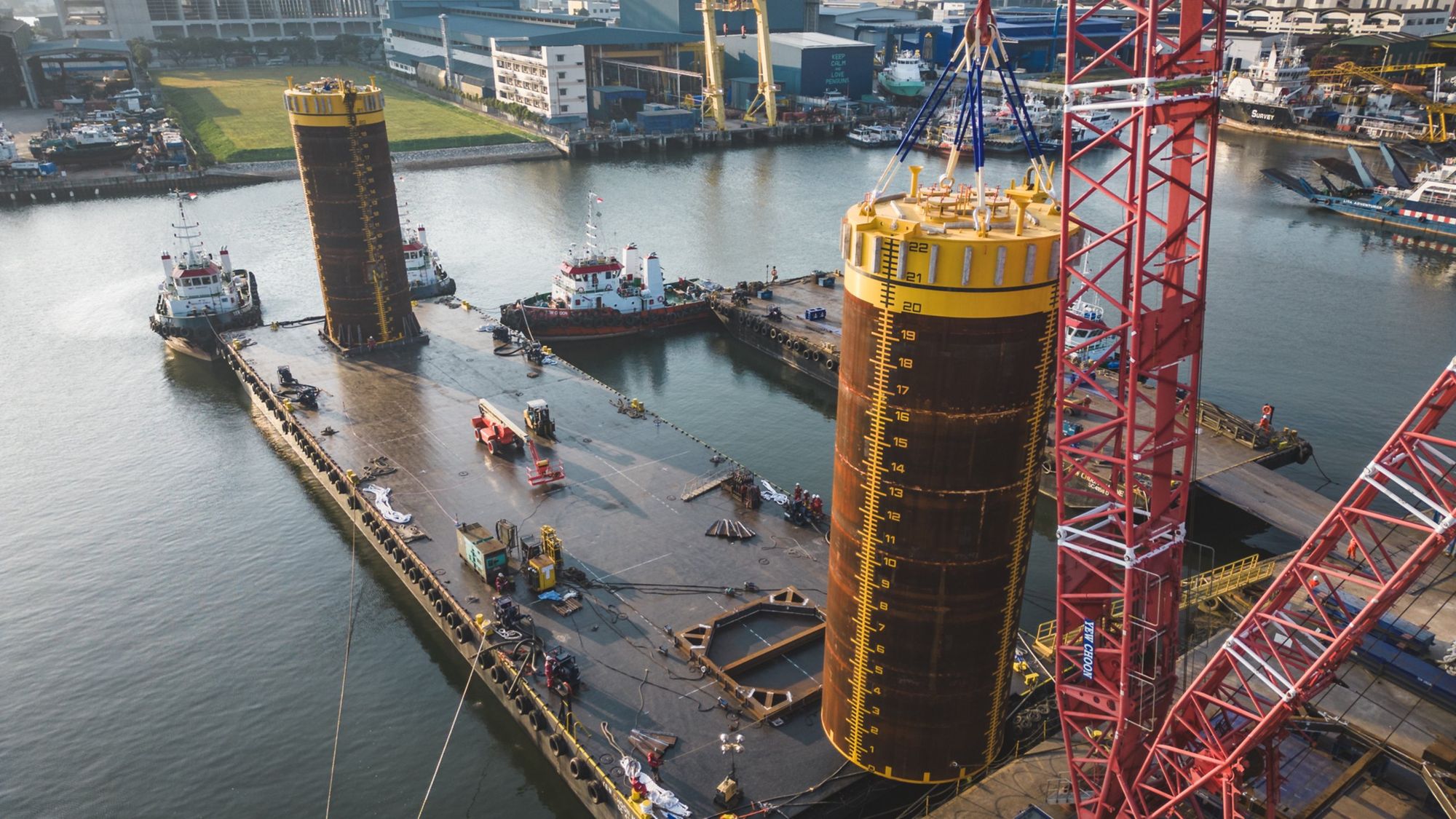 A pile lifted on NEXUS round slings by crawler crane beside barges and tugs at a waterfront fabrication yard
